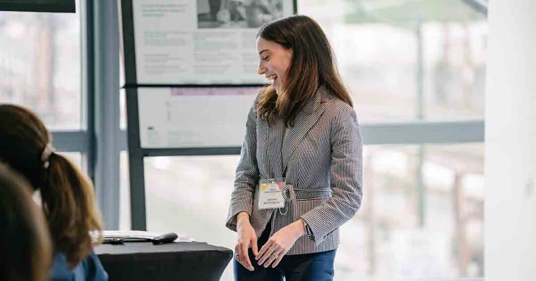 Allergy & Asthma Day Capitol Hill 2025: Leading the Charge Amid Federal Policy Shifts 35 A woman in a striped blazer with a conference badge smiles while standing at the front of a room, appearing to give a presentation. There are people seated nearby, and posters are displayed in the background.