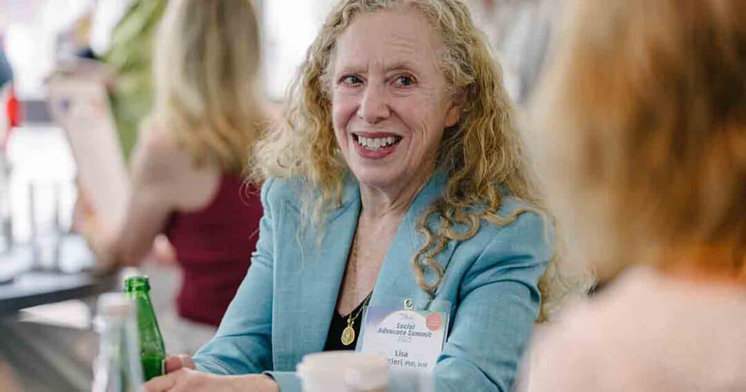 Allergy & Asthma Day Capitol Hill 2025: Leading the Charge Amid Federal Policy Shifts 34 A woman with curly blonde hair wearing a blue blazer smiles while sitting at a table. She has a name tag and holds a green bottle. Other people are visible in the blurred background.