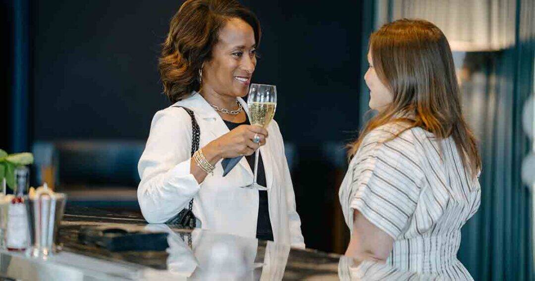 Allergy & Asthma Day Capitol Hill 2025: Leading the Charge Amid Federal Policy Shifts 33 Two women stand at a bar, conversing. One woman in a white blazer smiles and holds a glass of champagne, while the other, wearing a striped top, listens attentively. The setting appears relaxed and upscale.