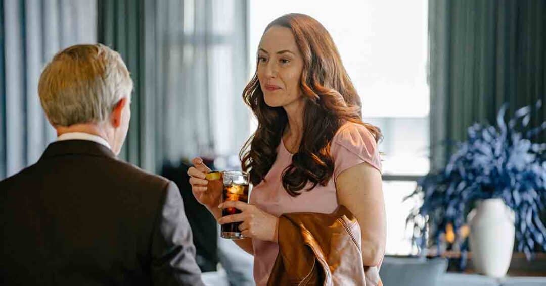 Allergy & Asthma Day Capitol Hill 2025: Leading the Charge Amid Federal Policy Shifts 32 A woman holding a drink and a brown jacket talks to a man in a suit inside a room with large windows and blue curtains. A leafy plant is visible in the background.