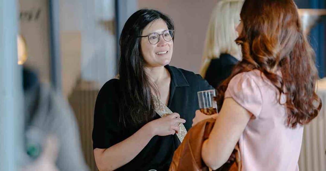 Allergy & Asthma Day Capitol Hill 2025: Leading the Charge Amid Federal Policy Shifts 30 Two women stand facing each other and talk. One woman with glasses is smiling, holding a drink. The other woman, with long red hair, is seen from behind, holding a coat. The background is out of focus.