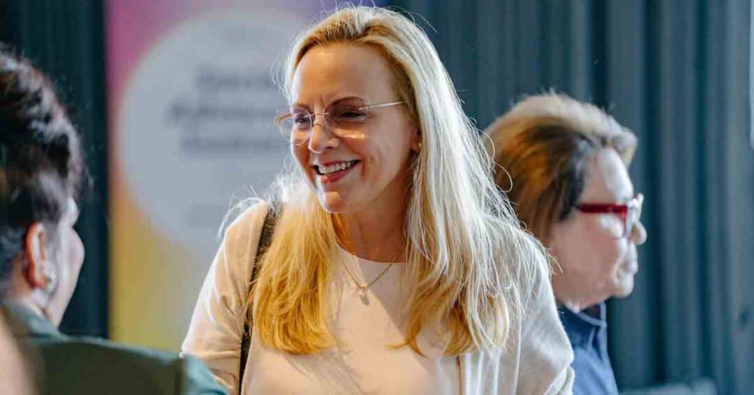 Allergy & Asthma Day Capitol Hill 2025: Leading the Charge Amid Federal Policy Shifts 28 A woman with long blonde hair and glasses smiles while talking to others indoors. Two other people are partially visible in the background. The setting appears casual and friendly.