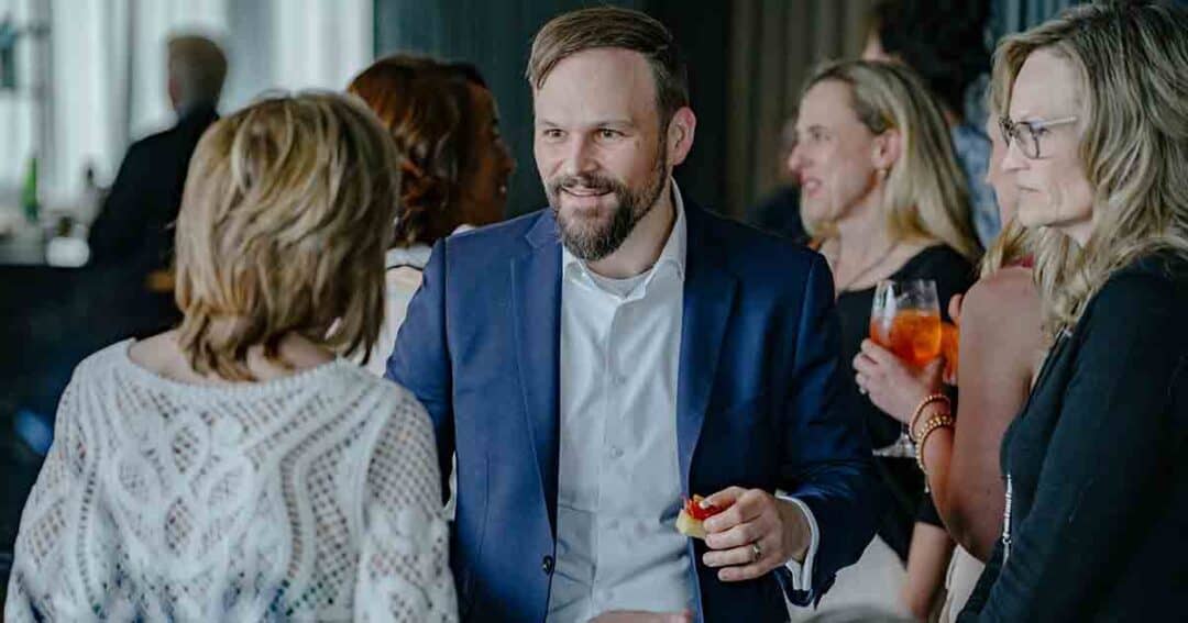 Allergy & Asthma Day Capitol Hill 2025: Leading the Charge Amid Federal Policy Shifts 27 A man in a blue suit smiles and talks with a group of women at a social gathering; he is holding a small appetizer, and people around him are holding drinks.