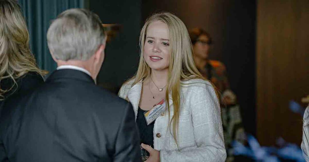 Allergy & Asthma Day Capitol Hill 2025: Leading the Charge Amid Federal Policy Shifts 26 A young woman with long blonde hair, wearing a white jacket and name tag, is speaking to an older man in a suit at an indoor event. Other people are blurred in the background.