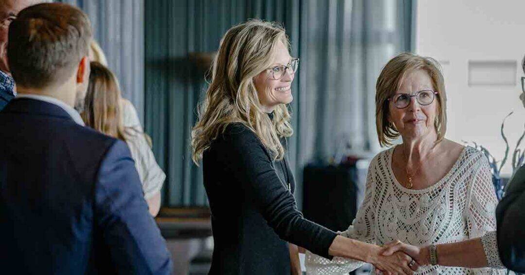 Allergy & Asthma Day Capitol Hill 2025: Leading the Charge Amid Federal Policy Shifts 23 Two women smiling and shaking hands at an indoor gathering, surrounded by other people in business or semi-formal attire. The setting appears to be a professional or social event.