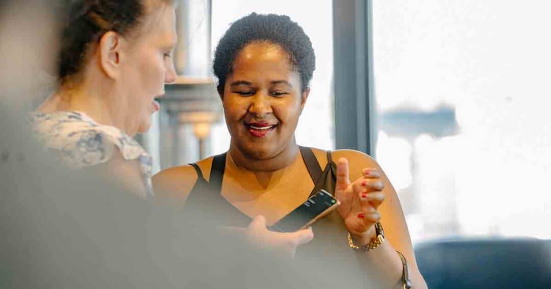 Allergy & Asthma Day Capitol Hill 2025: Leading the Charge Amid Federal Policy Shifts 19 Two women smiling and looking at a smartphone together indoors, one holding the phone while the other gestures, with natural light coming through the window behind them.