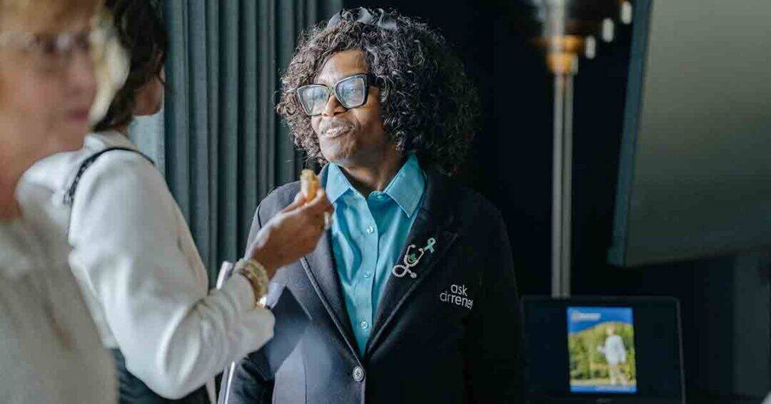 Allergy & Asthma Day Capitol Hill 2025: Leading the Charge Amid Federal Policy Shifts 18 A woman with curly hair and glasses, wearing a blue shirt and a dark jacket with pins, smiles while talking to another person at an indoor event. A screen is visible in the background.