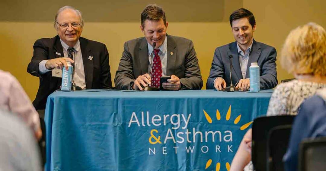 Allergy & Asthma Day Capitol Hill 2025: Leading the Charge Amid Federal Policy Shifts 58 Three men in suits sit behind a table with an "Allergy & Asthma Network" tablecloth, speaking and smiling at an event, facing an audience. Bottles of water and microphones are on the table.