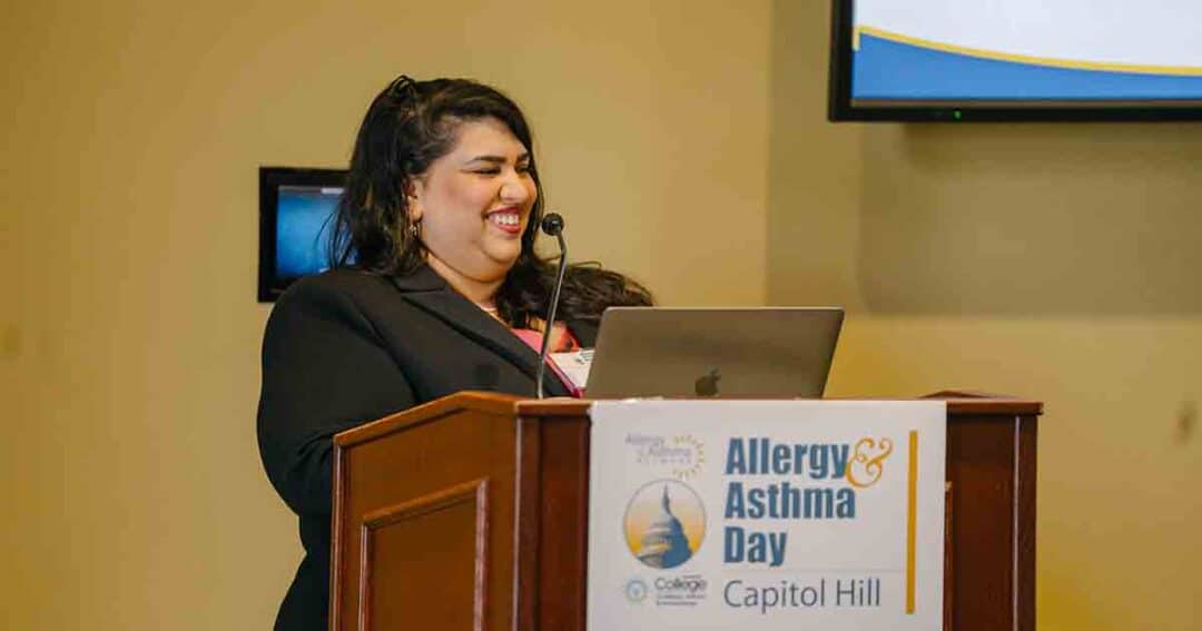 Allergy & Asthma Day Capitol Hill 2025: Leading the Charge Amid Federal Policy Shifts 15 A woman smiling stands at a podium with a laptop, speaking at an event labeled "Allergy & Asthma Day Capitol Hill." A presentation screen is partially visible in the background.