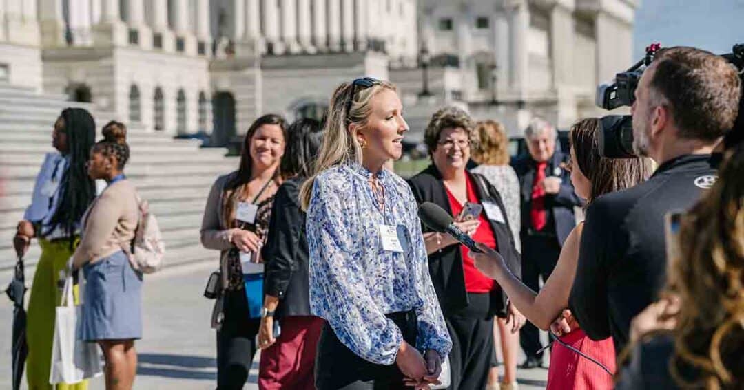 Allergy & Asthma Day Capitol Hill 2025: Leading the Charge Amid Federal Policy Shifts 11 A woman is being interviewed with a microphone by a reporter in front of a government building, with a group of people standing and talking in the background on the steps.