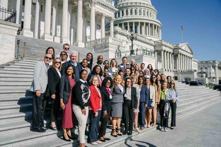 A large, diverse group of people pose for a photo on the steps outside the U.S. Capitol building under a clear blue sky. The group is smiling and dressed in professional attire.