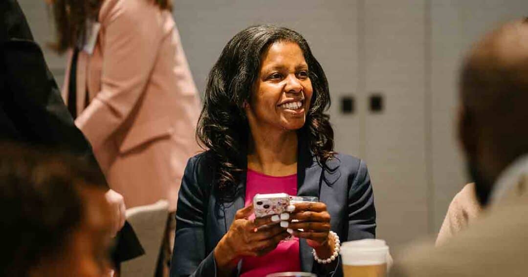 Allergy & Asthma Day Capitol Hill 2025: Leading the Charge Amid Federal Policy Shifts 21 A woman in a navy blazer and pink top smiles while holding a smartphone, seated at a table with a coffee cup. Other people are visible in the background, suggesting a social or professional event.