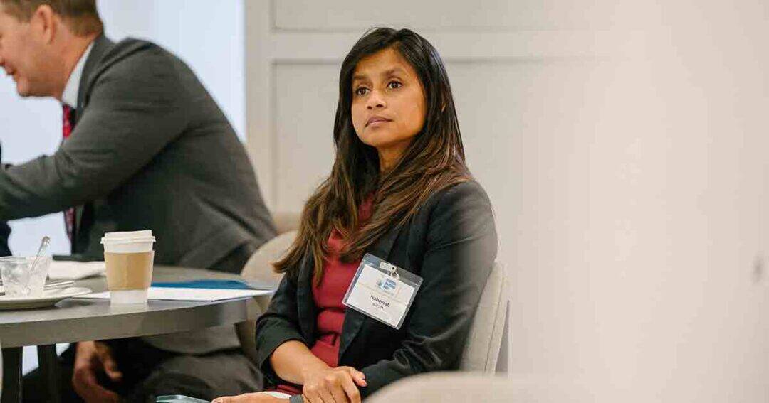 Allergy & Asthma Day Capitol Hill 2025: Leading the Charge Amid Federal Policy Shifts 20 A woman with long brown hair, wearing a dark blazer and red top, sits at a table during a meeting, looking attentive. A coffee cup and papers are on the table, with blurred people in the background.