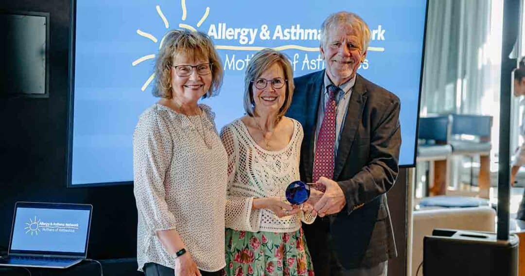 Allergy & Asthma Day Capitol Hill 2025: Leading the Charge Amid Federal Policy Shifts 37 Three smiling adults stand together indoors; one woman in the center holds a blue glass award. A screen behind them displays "Allergy & Asthma Network." A laptop with the same logo sits on a table nearby.