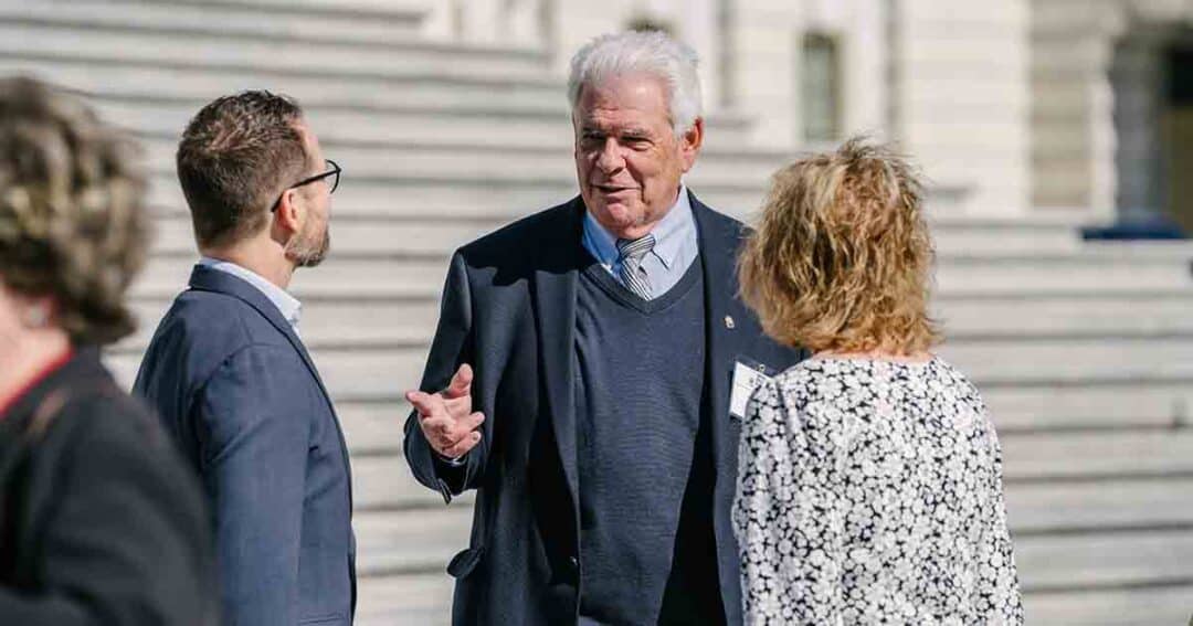 Allergy & Asthma Day Capitol Hill 2025: Leading the Charge Amid Federal Policy Shifts 7 Three people stand outdoors on stone steps, engaged in conversation. The man in the center gestures while speaking, wearing a suit and sweater; the others listen attentively. The background is out of focus.