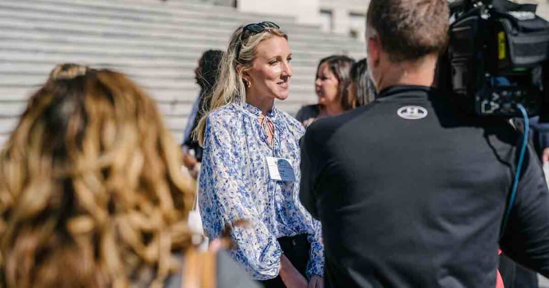 Allergy & Asthma Day Capitol Hill 2025: Leading the Charge Amid Federal Policy Shifts 9 A woman wearing a blue floral blouse and sunglasses on her head is being interviewed by a man with a video camera. They are outside, with blurred people and stone steps in the background.