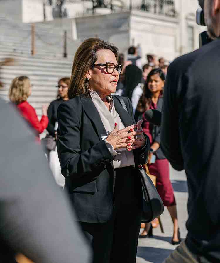 Allergy & Asthma Day Capitol Hill 2025: Leading the Charge Amid Federal Policy Shifts 5 A woman in a black suit and glasses speaks to reporters outside on marble steps, with microphones pointed at her and several people standing in the background.