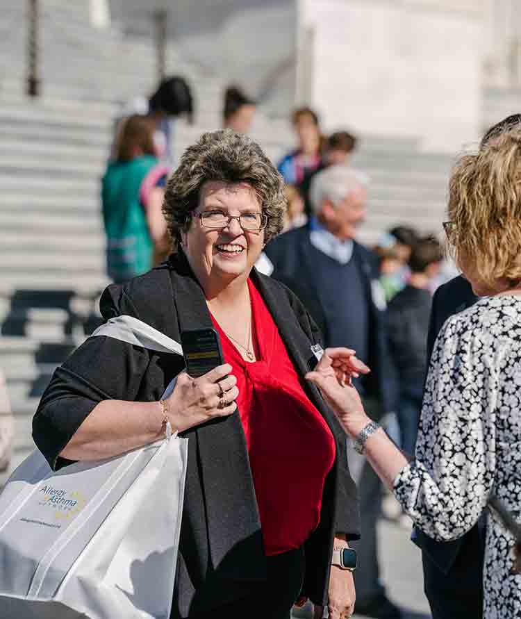 Allergy & Asthma Day Capitol Hill 2025: Leading the Charge Amid Federal Policy Shifts 8 A woman in a red shirt and black jacket smiles while holding a microphone and a tote bag, speaking to another person outdoors, with people and steps blurred in the background.