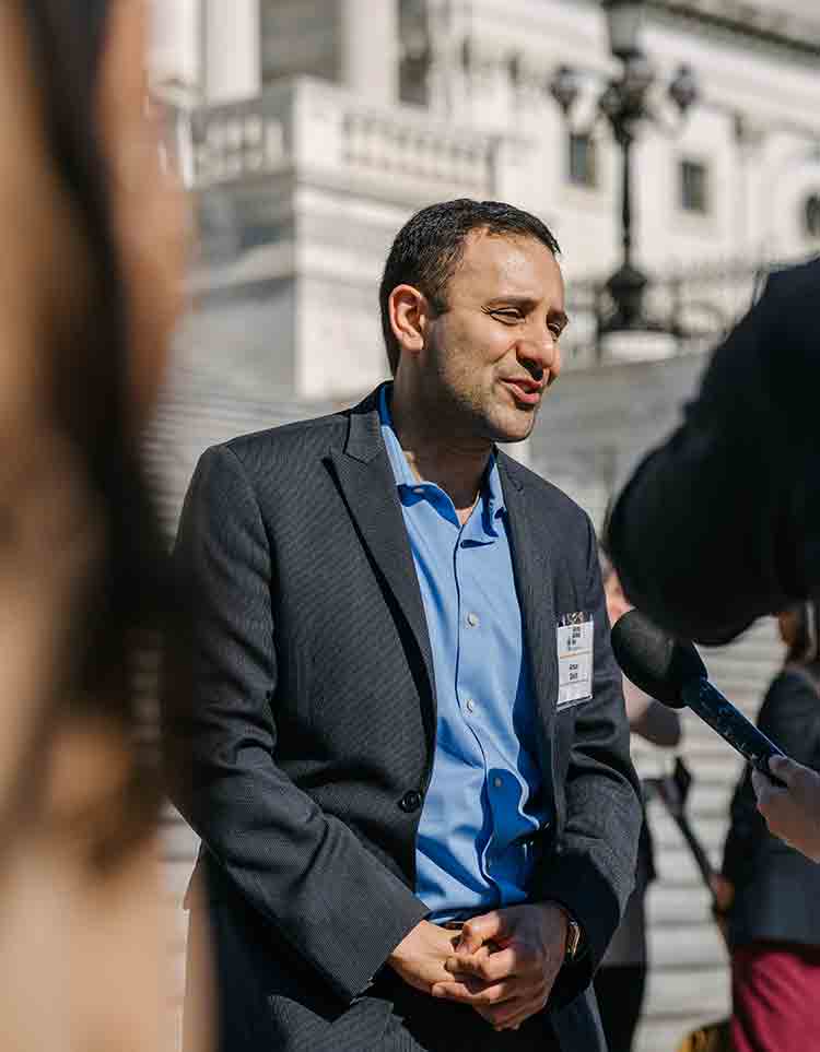 Allergy & Asthma Day Capitol Hill 2025: Leading the Charge Amid Federal Policy Shifts 10 A man in a blue shirt and dark blazer speaks to reporters outside a large government building, appearing engaged in conversation with microphones pointed toward him.