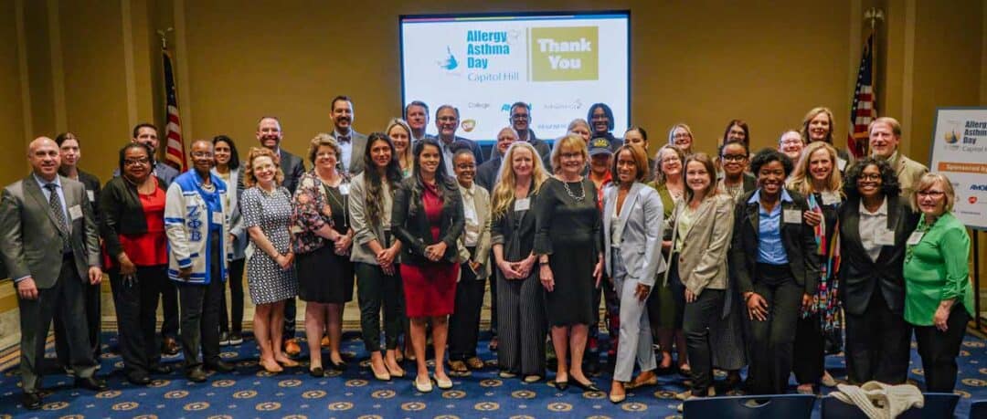 A large group of people, dressed in business attire, pose together in a conference room. Behind them is a screen displaying "Allergy Asthma Day Capitol Hill" and "Thank You." An American flag stands to the right.