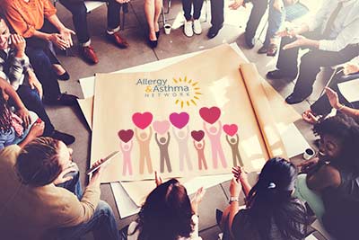 Ways to Give 8 A diverse group of people sit in a circle, discussing or collaborating. In the center, there's a large poster with the Allergy & Asthma Network logo and colorful figures holding heart shapes.