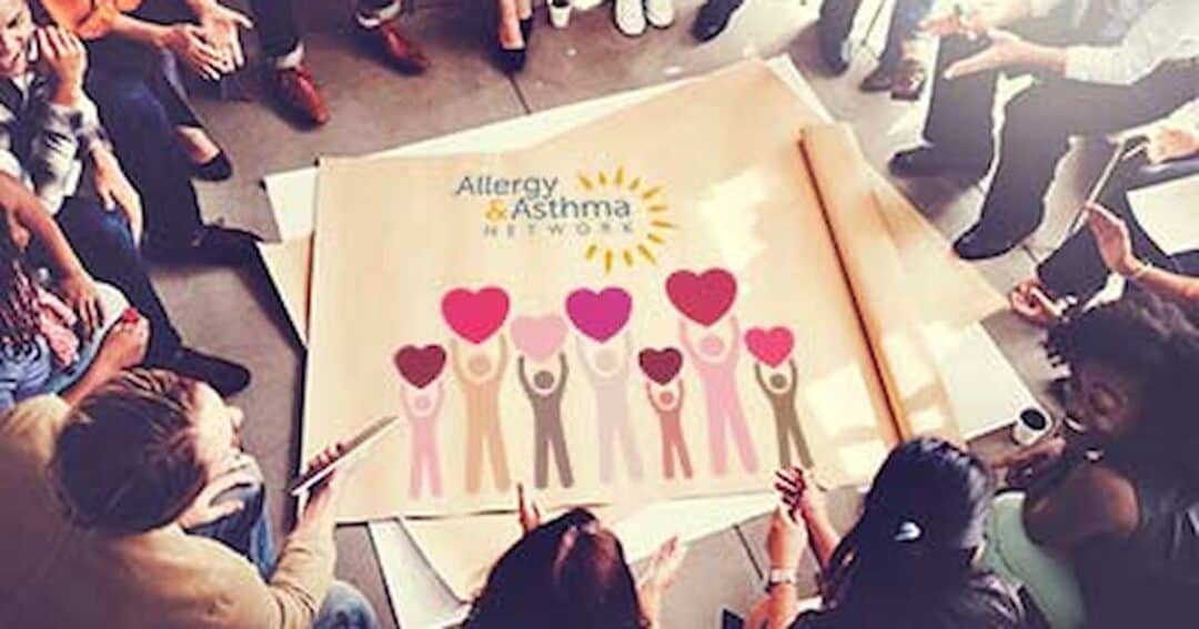 A group of people sit in a circle around a large paper with the "Allergy & Asthma Network" logo. The paper shows diverse figures holding up hearts. Everyone appears engaged, with some holding papers or pens.