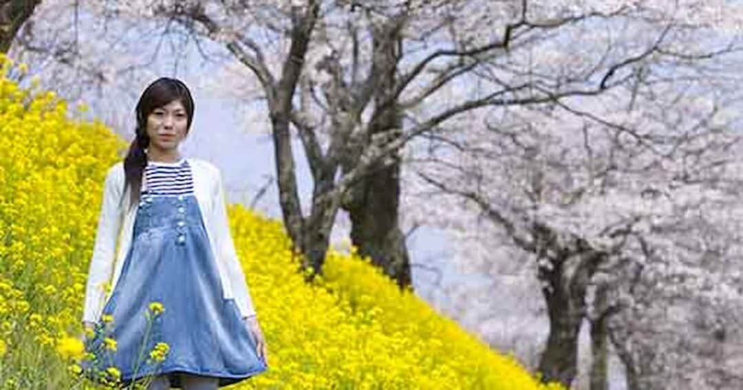Eye Allergies - Allergic Conjunctivitis 3 A woman in a denim dress and white cardigan stands among vibrant yellow flowers on a hillside. Behind her are blooming cherry blossom trees under a clear blue sky.
