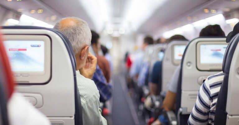 In the airplane cabin, passengers are seated, and from the back, a man with gray hair stands out. Seatback screens display content, contributing to the calm and well-lit atmosphere.