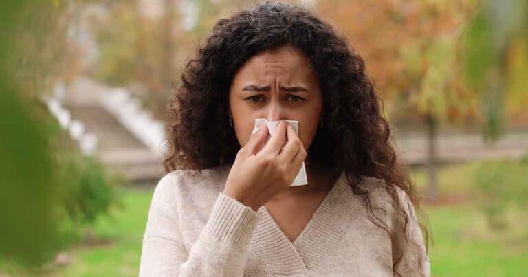 A person with curly hair is outdoors, holding a tissue to their nose. They appear to be experiencing allergies or a cold. Trees with autumn leaves are visible in the background.