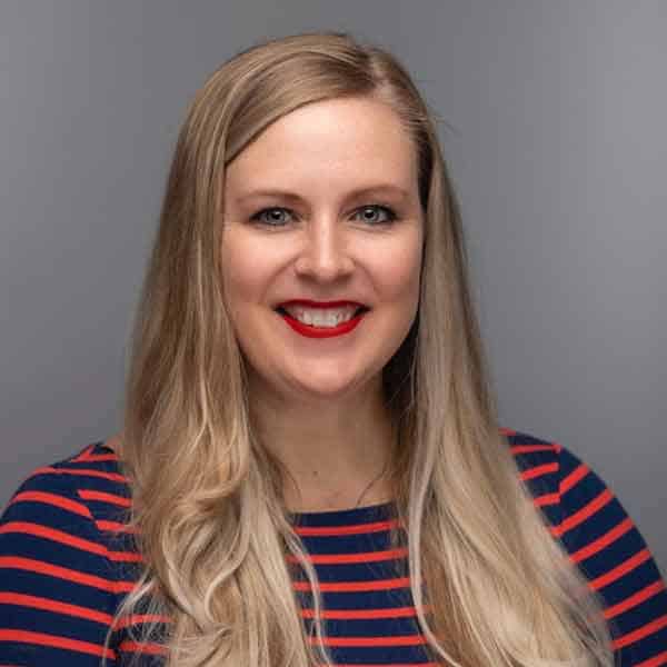 A woman with long blond hair and red lipstick smiles at the camera. She is wearing a dark blue top with red horizontal stripes, set against a plain gray background.