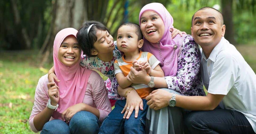 A joyful family sits together on grass in a park. Two women in pink headscarves, a girl kissing a toddler&rsquo;s cheek, and a smiling man all pose closely, exhibiting a sense of togetherness and happiness amidst lush greenery.