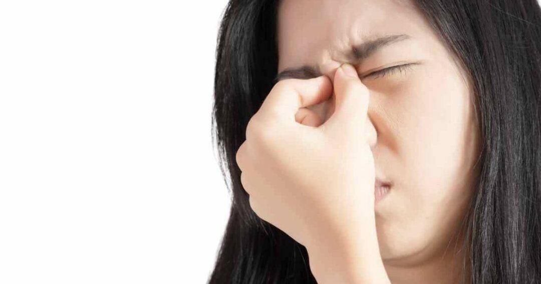A person with long dark hair pinching the bridge of their nose, eyes closed, as if experiencing discomfort or a headache, against a plain white background.