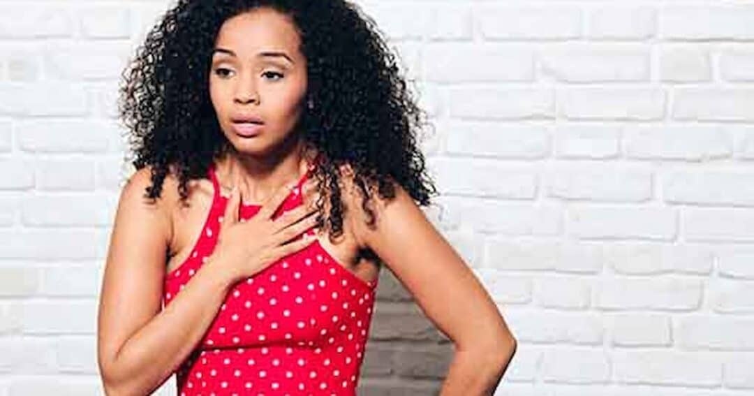A woman with curly hair and a red polka dot dress stands against a white brick wall. She has a hand on her chest and an expression of surprise or concern.