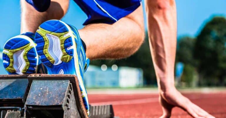Close-up of an athlete in starting blocks on a track. The focus is on the runner's blue and green sneakers and muscular legs, with a blurred background of a stadium under a clear blue sky.
