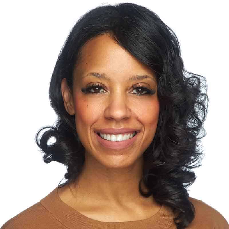 A woman with curly dark hair and brown skin smiles at the camera. She is wearing a light brown top and has natural makeup, with a white background behind her.