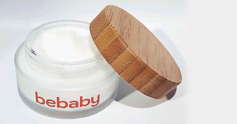 A glass jar of skincare cream with a wooden lid partially open, labeled "bebaby," is displayed on a light surface. The cream inside is white and smooth, and the lid has a natural wood grain texture.