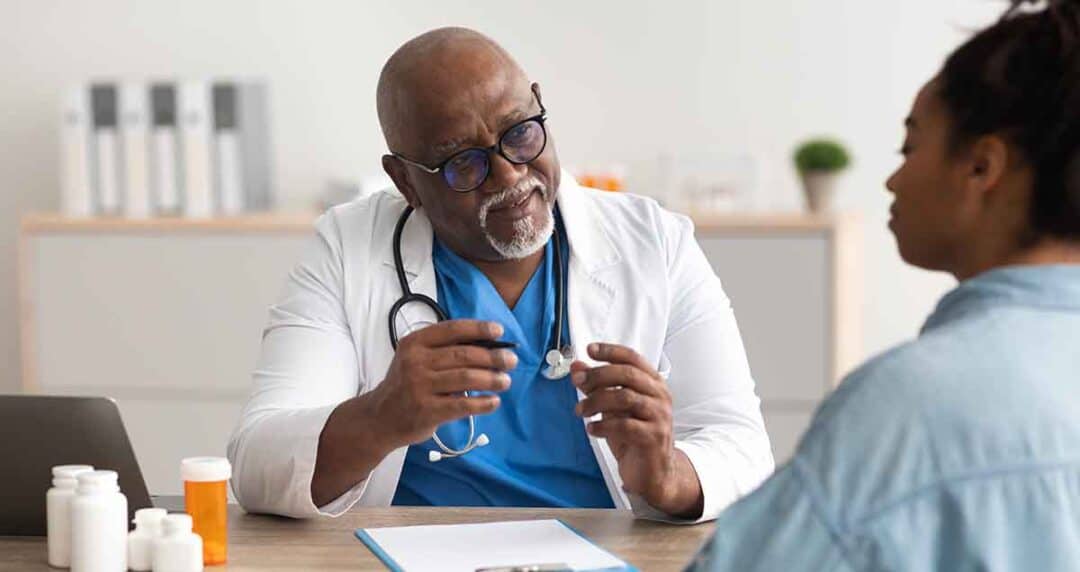 A doctor wearing glasses and a white coat sits at a desk with a stethoscope around his neck, holding a medical instrument. He talks to a patient who has curly hair and is wearing a denim shirt, bottles and a laptop are on the desk.