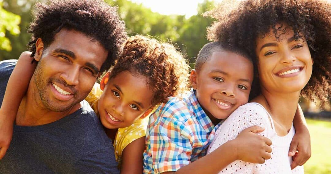 A happy family of four smiling outdoors. The parents are embracing their two children, a daughter and a son. They are all wearing casual clothing and appear content, with greenery visible in the background.