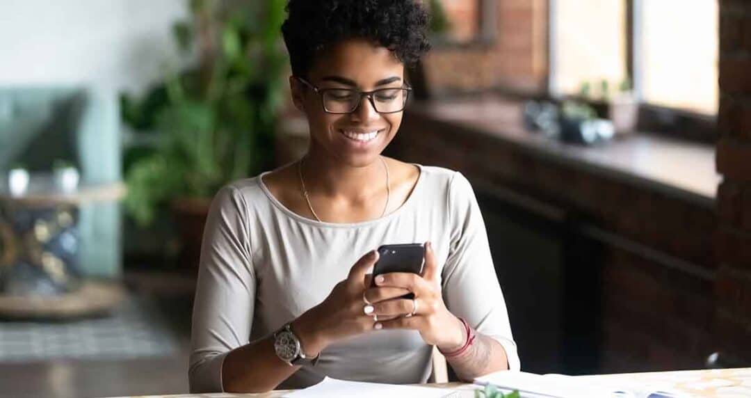 A smiling person with glasses sits at a table, looking at a smartphone. They are wearing a gray top and are in a well-lit room with plants and a large window in the background. Papers are scattered on the table.