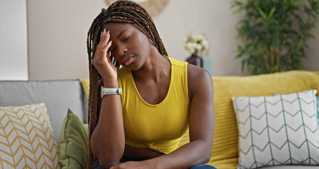A woman in a yellow top sits on a couch, looking pensive with her hand on her forehead. The room has patterned cushions and a potted plant in the background.