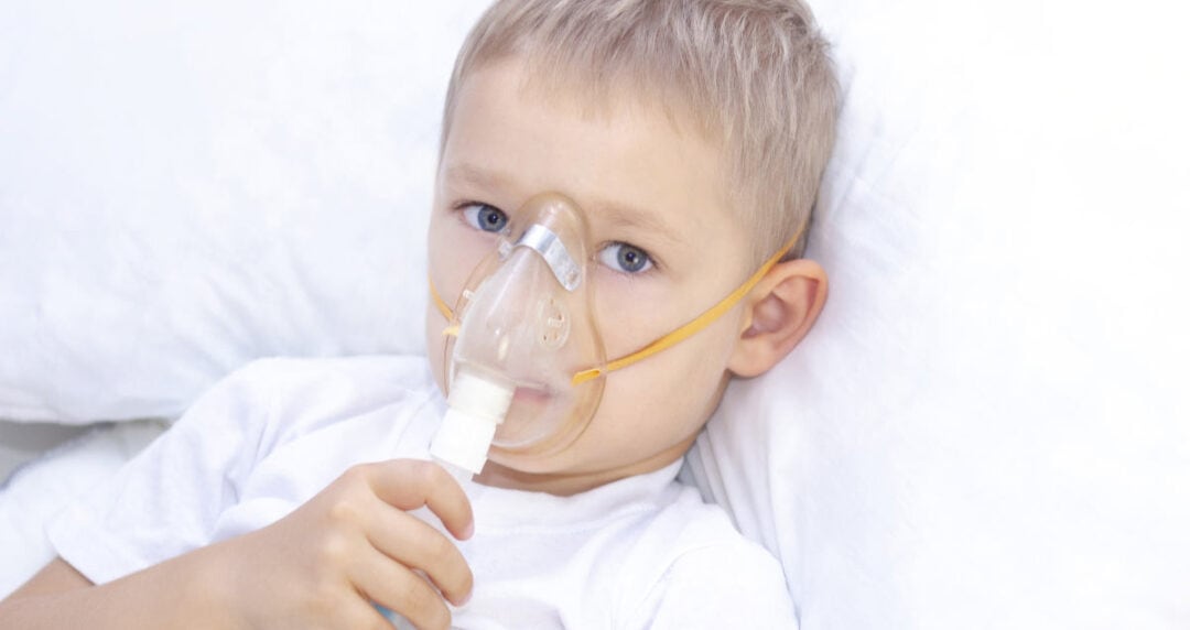 A young child with short blond hair lies in bed, wearing a white shirt. They are using a nebulizer mask connected to a device, suggesting they are receiving treatment. The background is a plain white pillow.