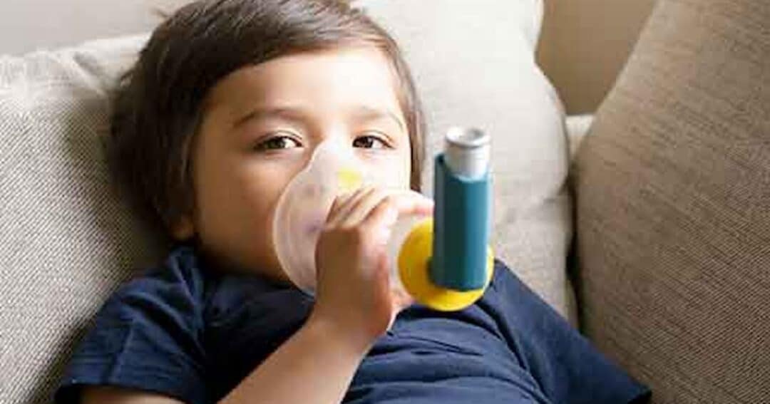 A child lies on a couch using an inhaler with a mask. The child has short brown hair and is wearing a dark blue shirt. The inhaler is blue with a yellow attachment, and the scene appears to be indoors with neutral-toned furniture.