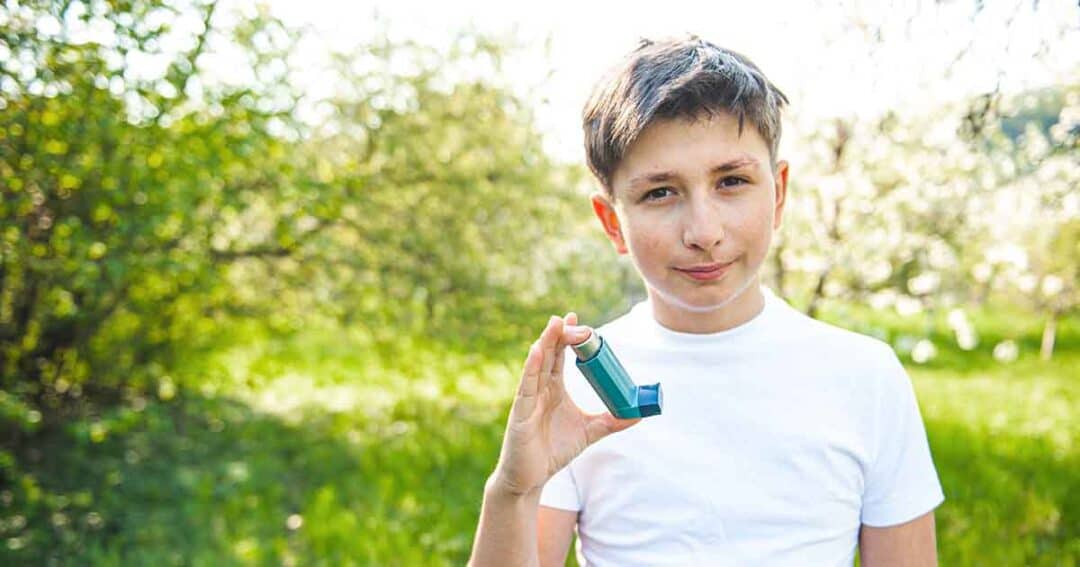 A boy standing outdoors in a green, sunny setting holds up an asthma inhaler while wearing a white shirt.