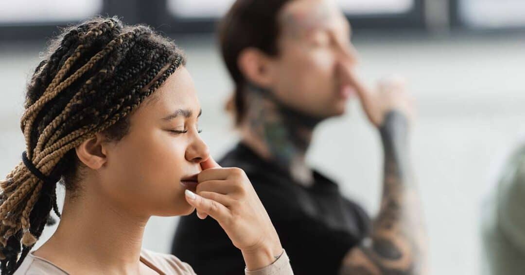 Alternative and Natural Asthma Treatments 2 Two people are practicing alternate nostril breathing. The person in the foreground has braided hair and appears focused, while the person in the background has tattoos and is also engaged in the breathing exercise.