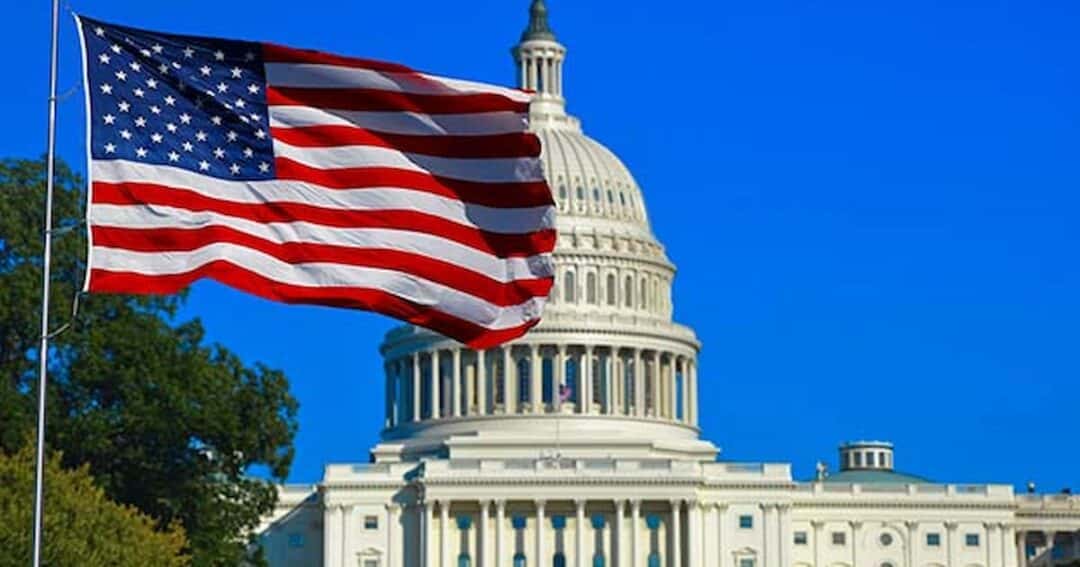 The image shows the United States Capitol building with a clear blue sky in the background. An American flag waves prominently in the foreground on the left side.