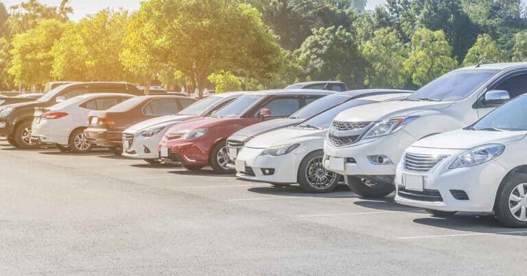 A row of various parked cars in an outdoor parking lot, with greenery and trees in the background on a sunny day.