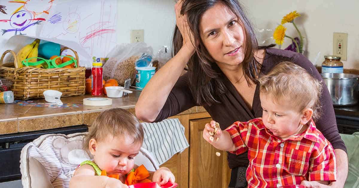 A woman sits at a kitchen counter with her hand on her head, looking tired. She is next to two toddlers, one playing with a red toy and the other holding food. The background includes colorful drawings and kitchen items.