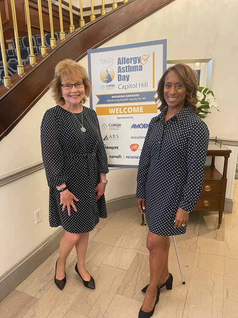 Two women in polka dot dresses stand smiling indoors at Capitol Hill's Allergy & Asthma Day. A welcome sign with sponsor logos is displayed in the background near a staircase.