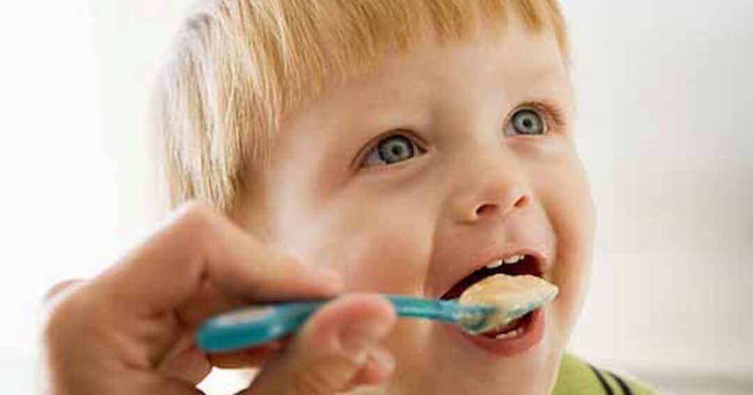 A young child with blonde hair is being fed with a blue spoon. The child looks happy and eager, with an open mouth ready to eat the food from the spoon. The background is softly blurred.