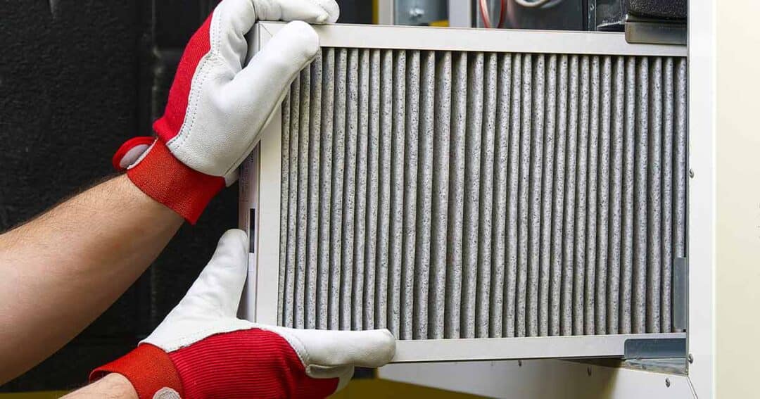 Indoor Air Quality and Indoor Air Pollution 4 A person wearing red and white gloves is installing or removing an air filter from a ventilation system. The filter appears to be slightly dirty, indicating use. The background shows parts of the ventilation unit.